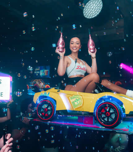 girl in a toy car above a nightclub crowd, holding two bottles with bubbles floating around